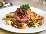 En gourmetret med en mediumstegt bøf toppet med karamelliserede løg og persille, serveret med ristede kartofler, gulerødder og pastinak og pyntet med hakkede krydderurter.