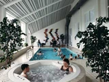 Folk slapper af i et indendørs poolområde; to sidder i et spabad i forgrunden, andre svømmer eller sidder ved poolen. Store planter og farverig vægkunst dekorerer det lyse, moderne rum.