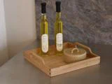 To flasker olivenolie og en dekorativ krukke med låg står på en træbakke, der er placeret på en grå bordplade.