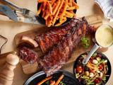 Grillede ribben på et træbræt med søde kartoffelpommes frites, en skål blandet salat og en lille gryde med cremet dippingsauce ved siden af.