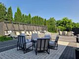Udendørs loungeområde med stribede, polstrede stole omkring små borde, liggestole under en pergola og træer i baggrunden på en solrig dag.