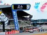 Bilerne holder i kø ved starten på 24-timers løbet i Le Mans, med tilskuere på tribunerne og rød, hvid og blå røg på himlen over et stort Michelin-skilt.