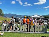 Stor udendørs folkemængde til en begivenhed med folk, der står bag et stort "#AUSTRIANGP"-skilt; bjerge, blå himmel og scene i baggrunden.
