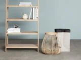 En træhylde med bøger og dekorationer står ved siden af en flettet kurv og en papirkurv mod en lysegrå væg og et betongulv.