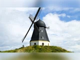 En traditionel sort-hvid vindmølle står på en græsklædt bakke under en delvis overskyet himmel med sine store vinger vinklet diagonalt.