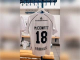 En hvid fodboldtrøje, der hænger i et omklædningsrum, med navnet McCoatt og tallet 18 trykt på ryggen.