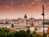 Den ungarske parlamentsbygning står ved siden af Donau-floden ved solnedgang, med dramatiske skyer på himlen og gadelamper, der indrammer bybilledet.
