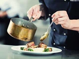 En kok i sort uniform anretter mad og hælder sauce fra en gryde over et fad med kød, kartofler og grønne grøntsager.