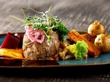 En gourmetret bestående af et rundt stykke kød toppet med mikrogrønt og syltede løg, serveret med ristede kartofler, gulerødder, romanesco-broccoli og en farverig grøntsagspuré.