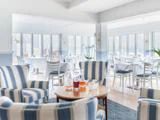 Lys, luftig restaurant med stribede lænestole og et rundt bord med glaskarafler; hvide borde og stole er arrangeret ved store vinduer i baggrunden.