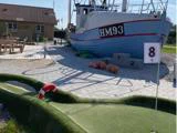 Et hul på en minigolfbane med en redningskrans ved siden af en stor blå og hvid båd mærket HM93, der er monteret på land, med bøjer og en gul bygning i nærheden.