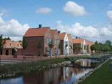 En række røde murstenshuse med sadeltag ligger ved siden af en kanal med en lille gangbro over vandet under en blå himmel med skyer.