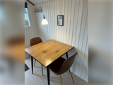Et lille spiseområde med et træbord, to brune stole, en pendel og minimalistisk vægkunst i form af et øje på de hvide panelvægge.
