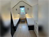 Et lille værelse med hvide paneler, hvor der står to enkeltsenge på hver sin side, et natbord af træ imellem dem, lamper over hver seng og et vindue på den bagerste væg.