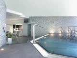Moderne indendørs swimmingpool med vægge af mosaikfliser, gelændere i rustfrit stål og et lille opholdsområde med stole og planter i baggrunden.