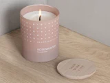 Et tændt SKANDINAVISK ROSENHAVE-duftlys i en pink beholder står på en træplade ved siden af det runde trælåg.