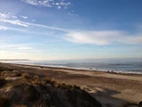 Græsklædte klitter i forgrunden, en sandstrand og blide bølger under en delvis overskyet blå himmel med nogle få mennesker, der går langs kysten.