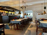 En hyggelig restaurant med træstole, borde dækket med grå duge, varme pendellamper og en bar i mørkt træ i hjørnet. Røde blomster pryder bordene.