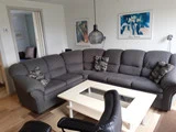 En moderne stue med en grå sektionssofa, mønstrede puder, et sofabord i hvidt glas, en sort stol og to indrammede kunstværker på væggen.