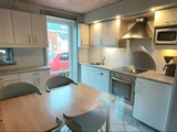 Moderne køkken med lyse træskabe, indbygget ovn, komfur med emhætte, mikroovn, kaffemaskine og et spisebord med fire stole. Et vindue viser en rød bil parkeret udenfor.