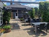 Udendørs terrasse med borde og stole under store Carlsberg-paraplyer, omgivet af grønt og blomster, på en solrig dag.
