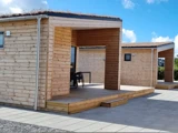 Moderne træhytter med fladt tag og små overdækkede verandaer, placeret på et asfalteret område under en blå himmel med enkelte skyer.