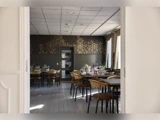 Elegant spisestue med dækkede borde og træstole, set gennem en åben døråbning; guldfarvet blomsterdesign på væggen og store vinduer med gardiner i baggrunden.