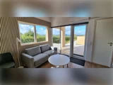 Moderne stue med grå sofa, rundt hvidt sofabord, stort vindue og glasdør med udsigt til en træterrasse og naturskøn udendørs udsigt med træer og blå himmel.