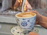 En person holder en blå og hvid mønstret kop latte med lattekunst foran en hyggelig pejs med ild i.