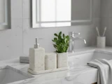 Et marmorbadeværelse med en sæbedispenser, en lille plante og en beholder på en bakke ved siden af en håndvask og et sammenfoldet håndklæde. Et spejl og en vandhane er også synlige.