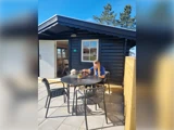 En kvinde sidder ved et sort udendørs bord med en hund i nærheden og spiser på en solrig terrasse foran en mørkeblå træhytte.