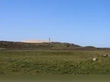 Et højt fyrtårn står på en sandbakke under en klar blå himmel, med en grøn græsmark og et hegn i forgrunden.