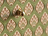 Nærbillede af en messingnål på et grønt og beige mønstret tapet med geometriske og blomstrede mønstre.