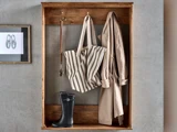 I et træhylde ved væggen ligger en stribet taske, en beige trenchcoat, en hundesnor og sorte gummistøvler ved siden af en indrammet skitse af en sko.