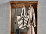 Et vægstativ af træ med kroge, hvor der hænger en stribet taske, en beige trenchcoat, en hundesnor og et par sorte gummistøvler på hylden nedenunder.
