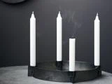 En rund, sort lysestage med fire hvide lys på en grå flade; det ene lys er slukket, og der stiger røg op fra vægen.
