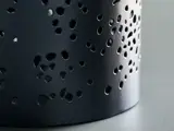 Nærbillede af en sort metalgenstand med mange ovale og runde huller, der sidder på en glat grå overflade.