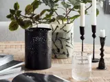 Et træbord med en sort udskåret lanterne, en mønstret vase med grønne bladstilke, tre lysestager med hvide stearinlys, et glas vand og åbne magasiner.