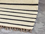 Et beige vævet tæppe med sorte vandrette striber og frynser i den ene kant, der ligger på et gråt, struktureret betongulv.