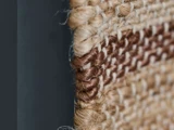 Nærbillede af kanten på et vævet tæppe eller en måtte, der viser beige og brune fibre, der er tæt flettet sammen på en mørk baggrund.