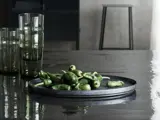 En metalbakke med friske grønne peberfrugter står på et blankt, mørkt bord ved siden af flere høje, røgfarvede glasbægre. En skammel og en sløret baggrund er synlige.