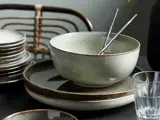 En keramikskål med to spisepinde i metal hviler på stablede tallerkener ved siden af en glaskop og flere tallerkener, med en stol i baggrunden.