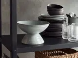 En grå keramikskål stablet på hovedet på en anden skål, ved siden af en bunke stablede tallerkener, skåle, glas og redskaber på en sort hylde med en flettet kurv under.
