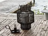 Et hvidt lys i en sort trådlygte og et andet lys i en sort metalholder står på et rustikt træbord ved en forvitret havn over vandet.