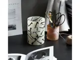 Et skrivebord med en hvid og sort vase med abstrakt mønster, en kop med sakse og blyanter, en spole med garn og åbne magasiner eller bøger.