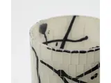 Nærbillede af en cylindrisk hvid vase med kakkelstruktur og abstrakte sorte streger og stænk som dekoration.