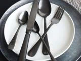 Et sæt matsort bestik med kniv, gaffel og to skeer arrangeret på en hvid tallerken med en mørkegrå oplader under.
