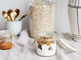 Et glas yoghurt med granola og frugt står på et bord. I nærheden står syltetøjsglas og havregryn, guldskeer i en kop og en hvid vase med sorte streger.