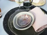 Et nærbillede af stablede keramikfade med en lilla mønstret skål, en beige serviet, et glas hvidvin og en sort vævet dækkeserviet på et bord.