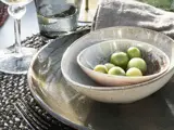Nærbillede af en borddækning med indlejrede keramikskåle, grønne oliven, en grå serviet, en tallerken, en gaffel og glas med vand og vin på en vævet dækkeserviet.